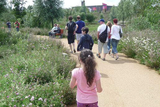 Families strolling on a park path, surrounded by flowering shrubs and greenery.